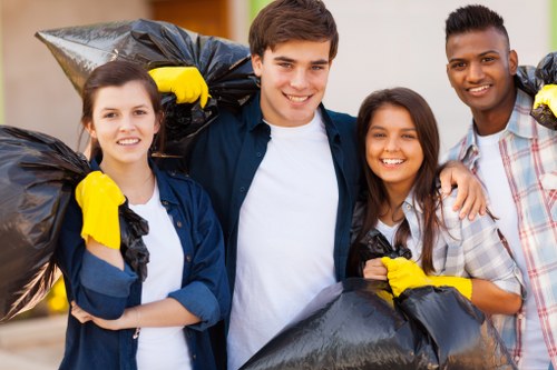 The image depicts four individuals standing outdoors on a paved driveway, each holding a large black plastic rubbish bag filled with waste. The group consists of two women and two men, all smiling and dressed casually; one woman and one man are wearing yellow cleaning gloves. The individuals are positioned closely together, with the background showing a residential area with blurred greenery and a house. The rubbish bags are clearly visible with some appearing slightly twisted or folded at the top, indicating they are filled and closed. The scene suggests a community or group effort in rubbish collection or clearance, aligning with professional waste removal services such as those offered by House Clearance Basildon, serving areas like Basildon and surrounding postcode districts. The lighting is natural and bright, highlighting their cheerful expressions and the black rubbish bags, emphasizing cleanliness and disposal activities characteristic of rubbish clearance projects.