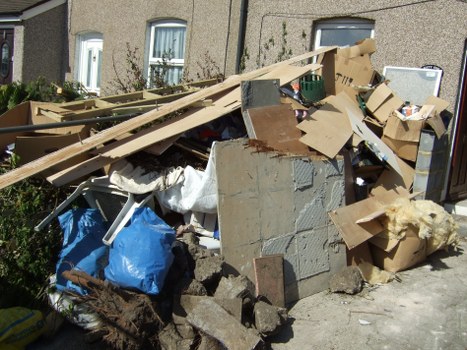 A large pile of mixed waste materials is situated outdoors on a paved surface next to a residential building. The rubbish includes broken wooden planks, cardboard boxes, and irregularly shaped concrete and brick fragments. Some cardboard boxes are partially flattened or torn, with pieces of packaging and paper visible. The wooden planks are weathered, with rough textures and light brown tones, some leaning against the pile. There are plastic bags, notably a blue one, among the debris, partially covered by a white cloth or tarp. The background shows a residential building with a beige exterior wall and windows, indicating this rubbish removal site is in a suburban area. The scene is naturally lit, casting minimal shadows, and the overall context suggests an ongoing clean-up or house clearance effort, relevant to services provided by House Clearance Basildon in Basildon, Essex.