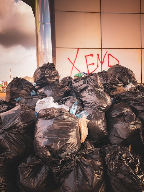 A large pile of black rubbish bags, some partially torn, stacked against a modern building with a tiled exterior wall. The bags are filled with household waste and are visibly bulging, with a few bott