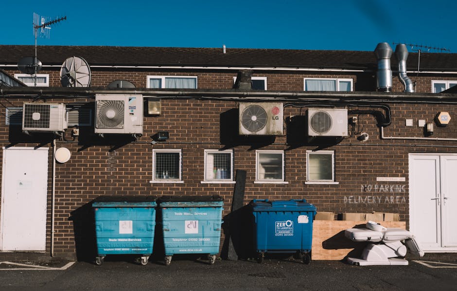 A large pile of black rubbish bags, some partially torn, stacked against a modern building with a tiled exterior wall. The bags are filled with household waste and are visibly bulging, with a few bott