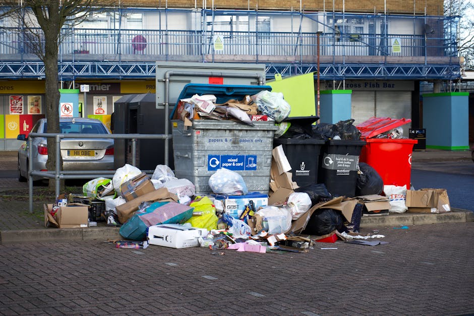 A narrow, urban alleyway cluttered with a large black garbage bag and a metal trolley filled with discarded waste, situated between graffiti-covered walls and exterior building facades. Behind the tro