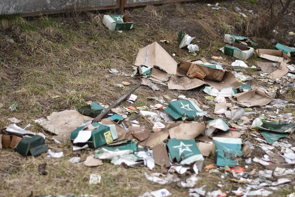 An outdoor scene showing a patch of uneven ground with sparse grass and dirt, strewn with discarded cardboard boxes, many of which are crushed or torn, and some are partially collapsed. The boxes are